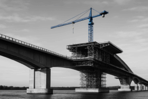Black-and-white photo of a bridge under construction with visible scaffolding, cranes, and exposed beams.